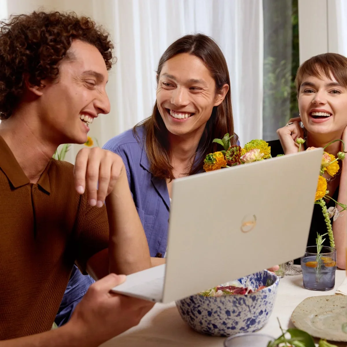 Three people sitting at a table looking at a laptop, smiling and laughing, with flowers on the table.