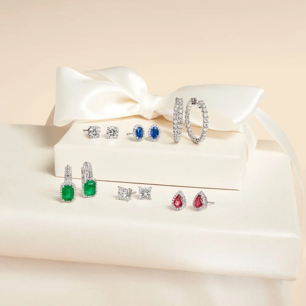 Collection of various diamond and gemstone earrings displayed on a cream-colored gift box with a satin ribbon
