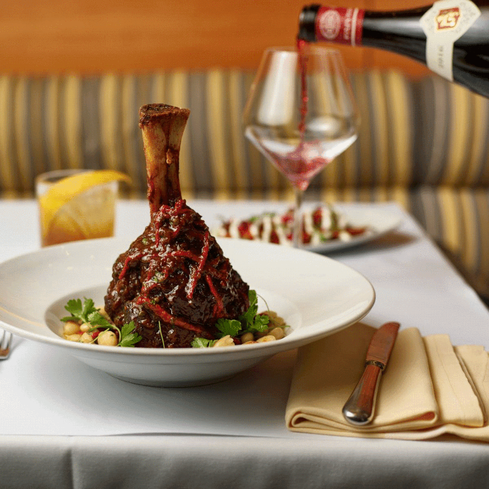 A plate with a cooked lamb shank covered in sauce, garnished with herbs, with a lemon wedge and a glass of red wine pouring from a bottle, set on a white tablecloth with a napkin and cutlery.