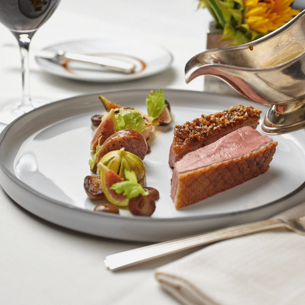 A plated dish featuring slices of duck breast with a crust, garnished with a sauces, and accompanied by vegetables and garnishes, with a glass of wine in the background.