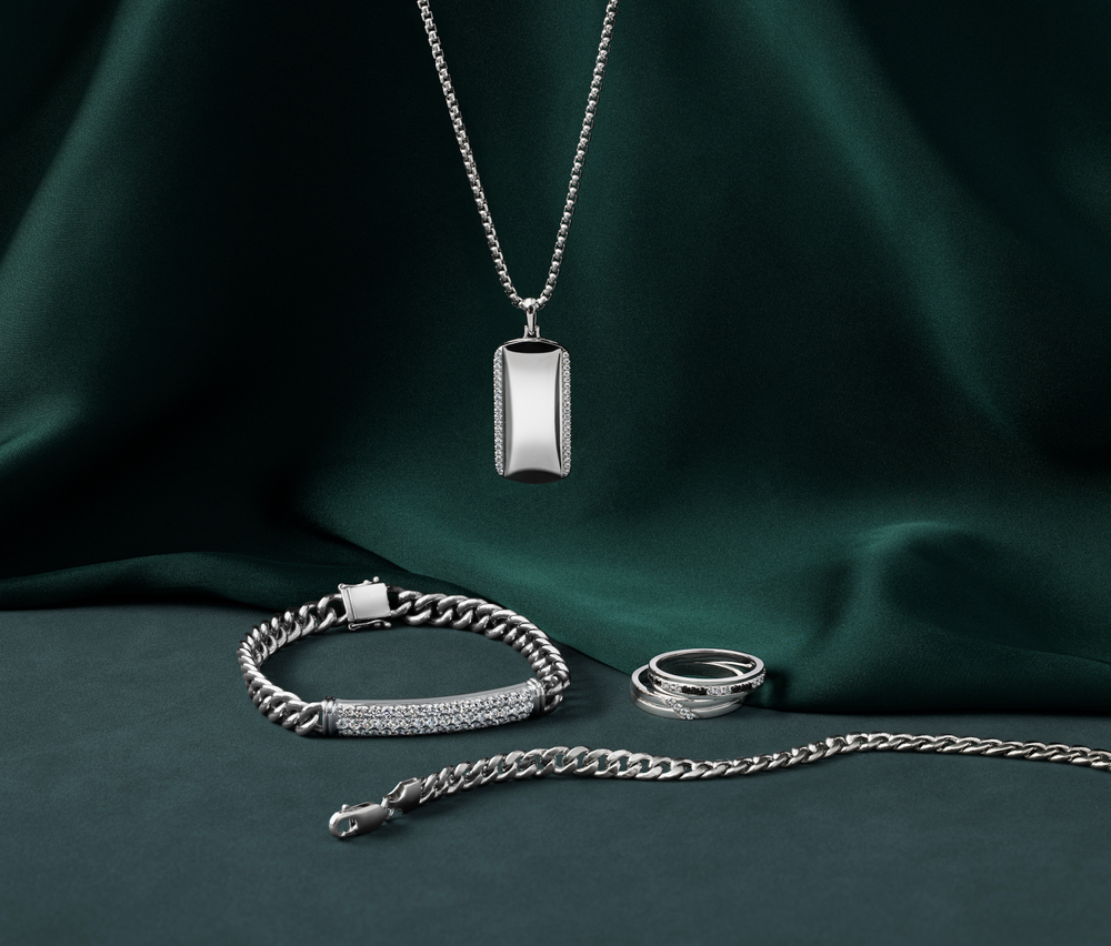 Silver jewelry set including a dog tag necklace, chain bracelet, ring, and chain necklace displayed on dark green fabric background.