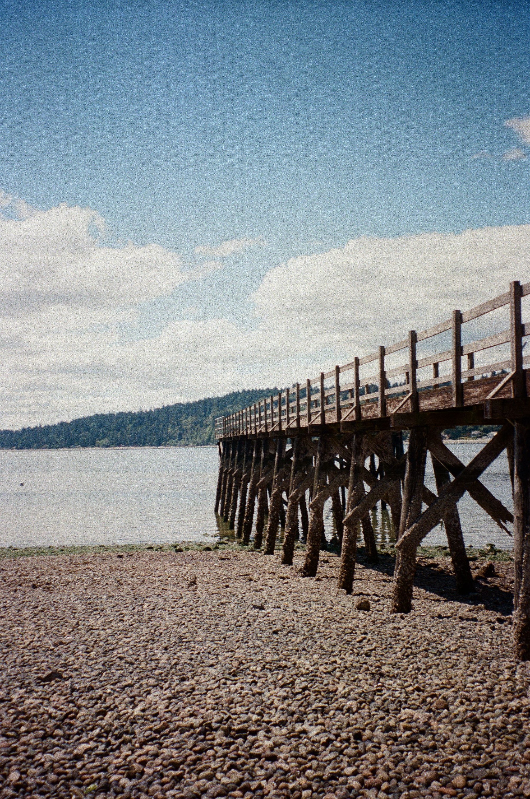 A wooden pier extends into a river with a rocky beach in the foreground and a forested hill in the distance, under a partly cloudy sky.