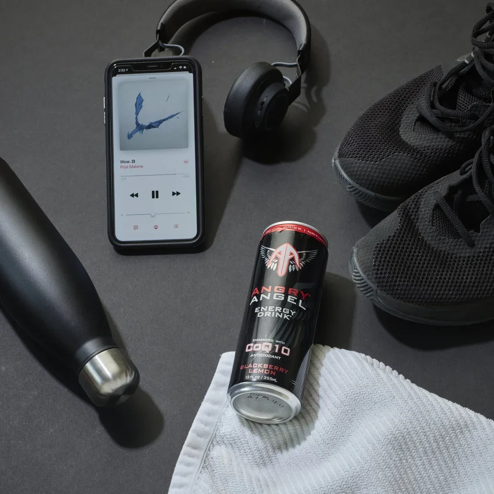 A gym workout setup including a smartphone playing music, wireless headphones, a water bottle, a can of Angry Angel energy drink, a white towel, a pair of black athletic shoes, and a black water bottle on a black surface.