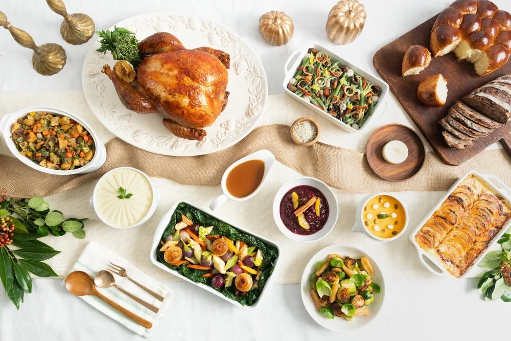 A Thanksgiving feast including a roasted turkey with stuffing, various salads, bread, and sides, on a white table with decorative pumpkins and greenery.
