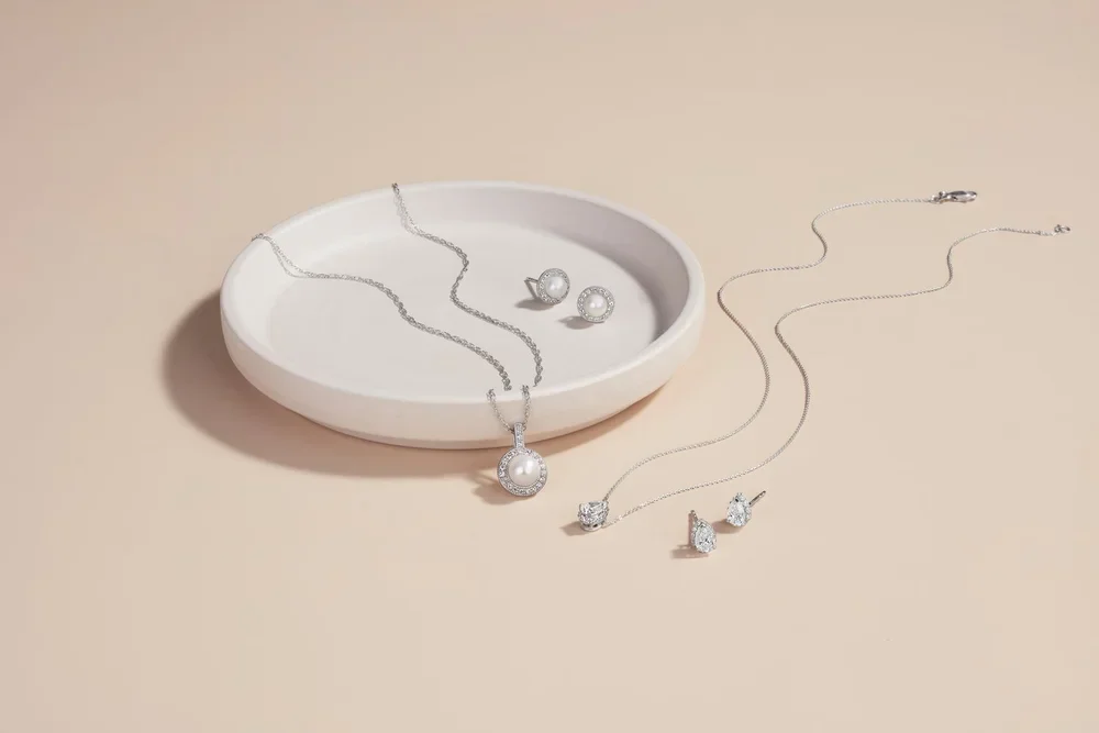 Jewelry set including a pearl necklace with a pendant, pearl earrings, and pairs of diamond earrings on a beige surface with a white dish.