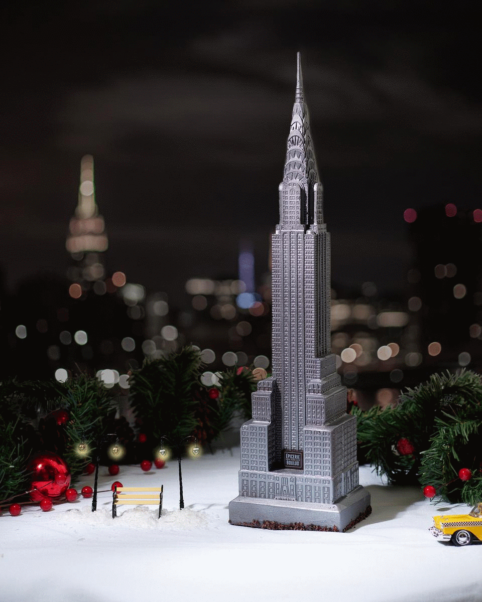 Miniature replica of the Empire State Building decorated with festive Christmas decorations, placed on a snowy surface with a blurred cityscape background at night.
