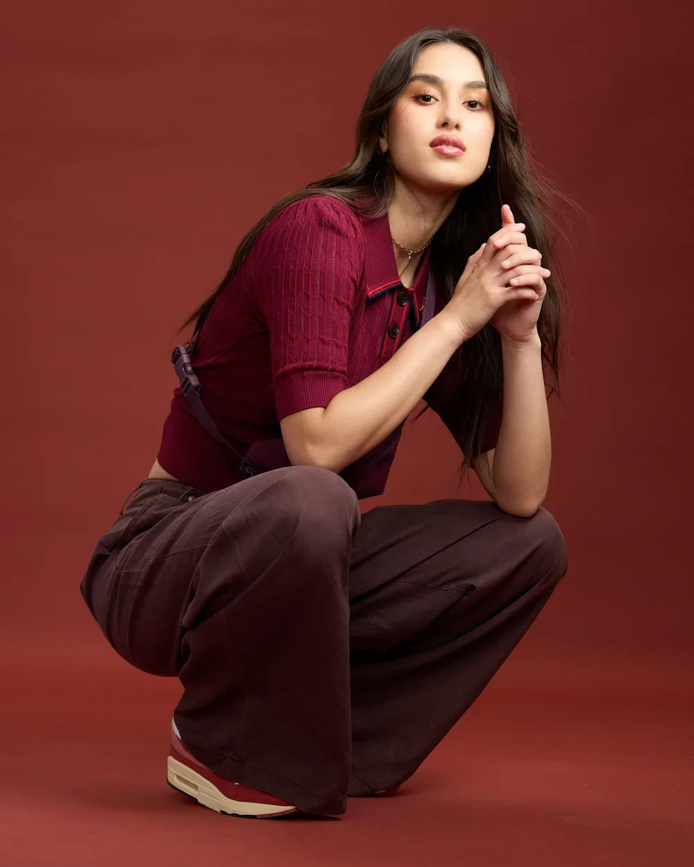 Young woman with long dark hair wearing a red shirt and brown pants, squatting against a red background.