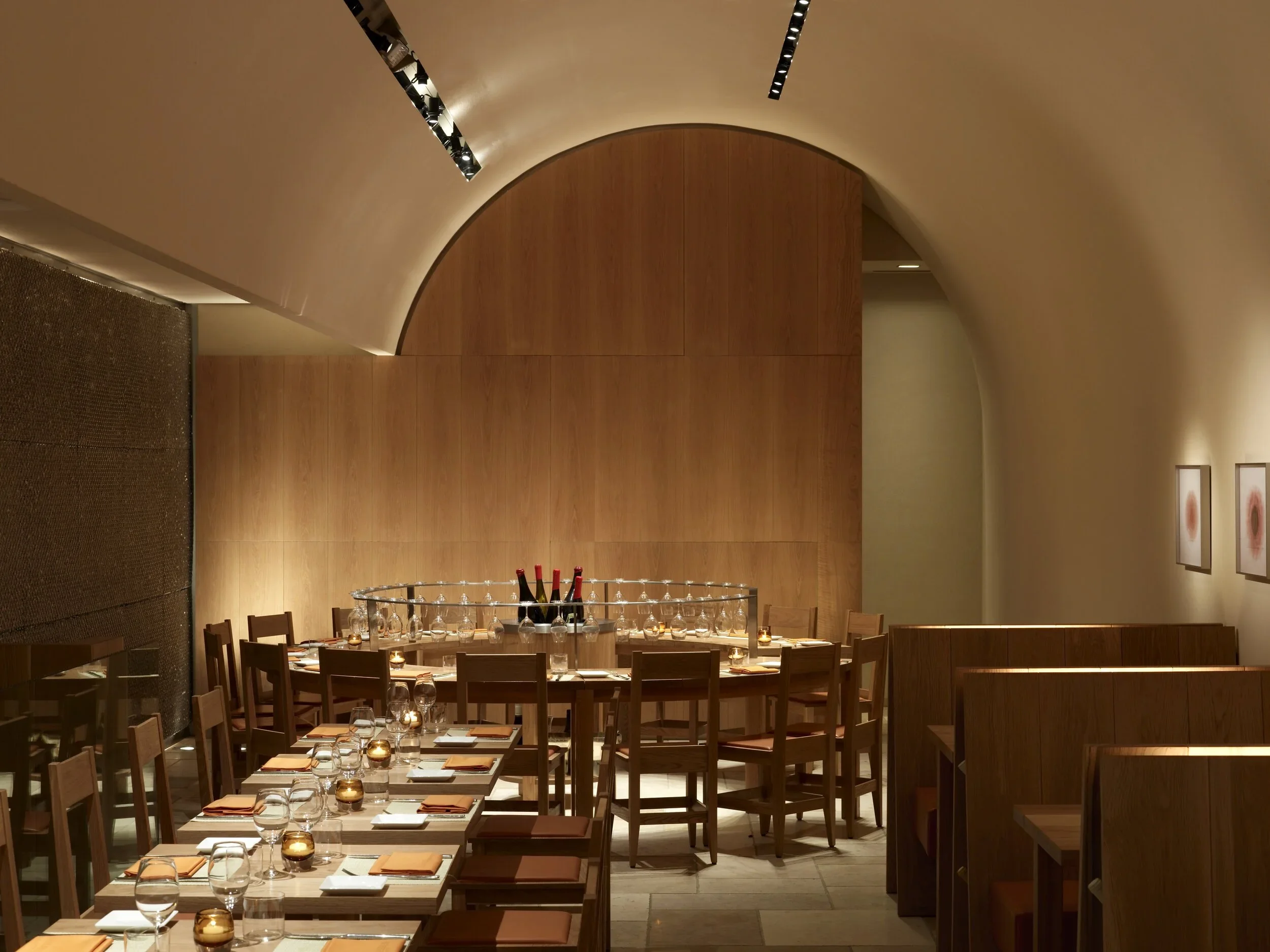 A modern restaurant interior with a large, curved wooden wall, tables set with wine glasses, plates, and orange napkins, warm ambient lighting, and framed artworks on the right wall.