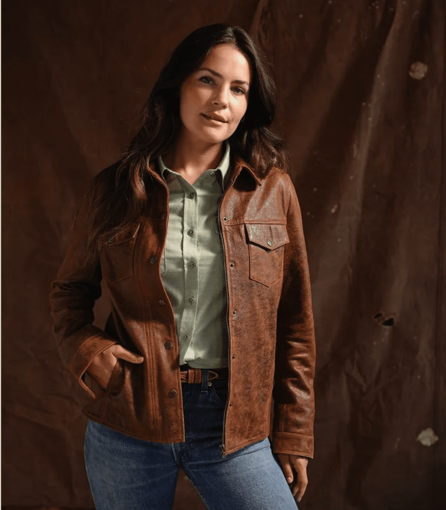 A woman with long dark hair wearing a light green shirt and a brown leather jacket standing against a brown textured background.