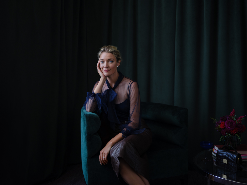 A woman seated on a teal velvet armchair in front of dark curtains, wearing a sheer navy blouse with a bow and a black skirt, with a side table holding a bouquet of flowers and a black purse.
