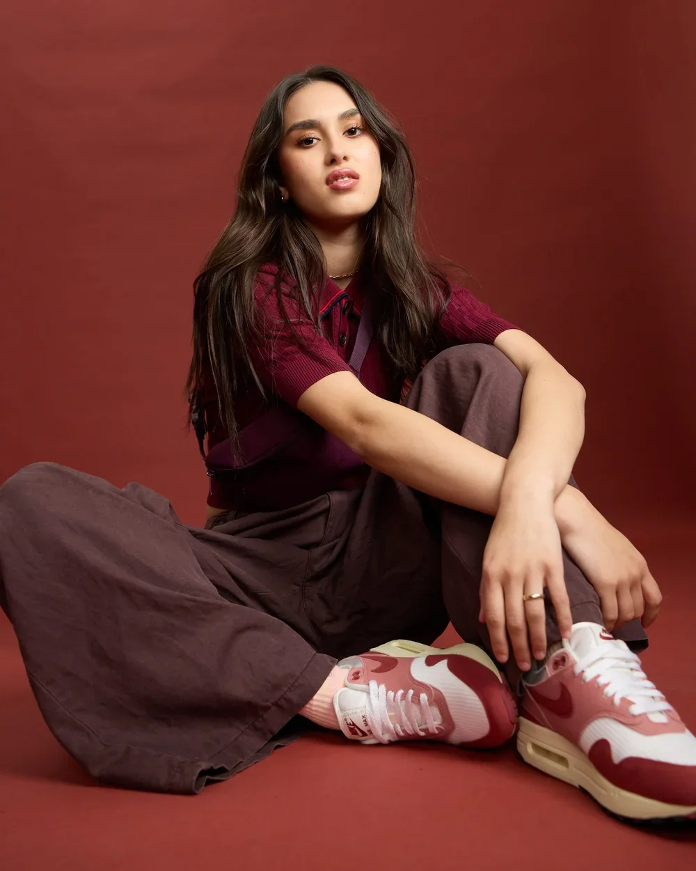 A young woman with long dark hair, wearing a maroon shirt, brown baggy pants, and pink and white sneakers, sits on the floor against a reddish-brown background with her legs crossed and arms resting on her knees.