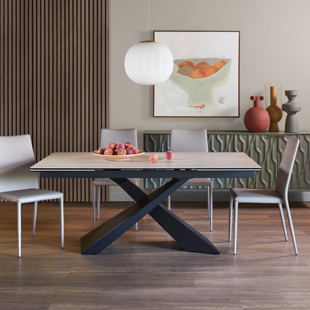 Modern dining room with a wooden table, surrounded by four white chairs, a bowl of peaches on the table, and a white pendant light overhead. The background features a beige wall with abstract art and a green textured sideboard with decorative vases.