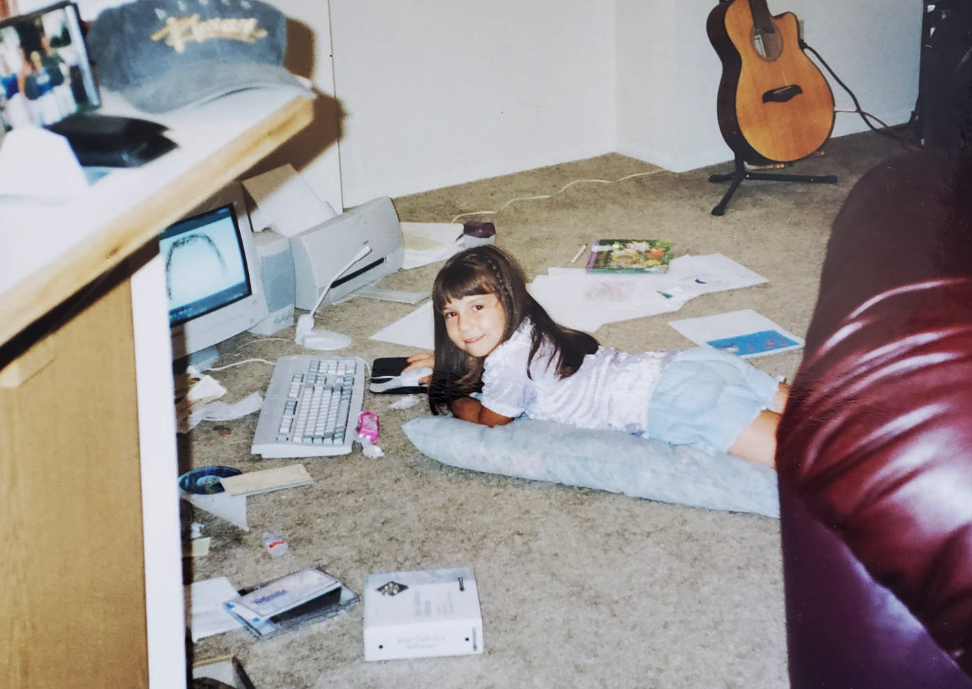 A young girl lying on the carpeted floor of a cluttered room, using a computer mouse next to a keyboard, with papers, books, and various items scattered around. An acoustic guitar stands in the corner, and a flat-screen TV is on a wooden shelf to the left.
