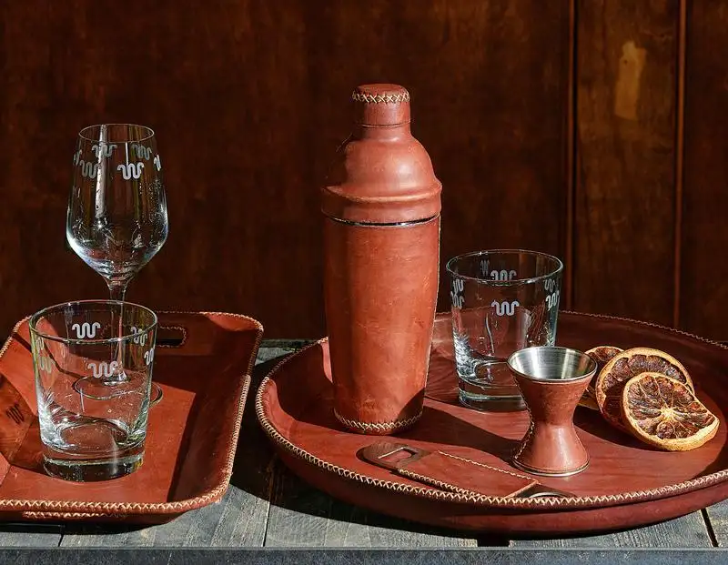 A still life arrangement on a wooden surface with two leather trays, a leather cocktail shaker, a copper jigger, three sliced dried oranges, and a wine glass and a tumbler with W-shaped patterns.