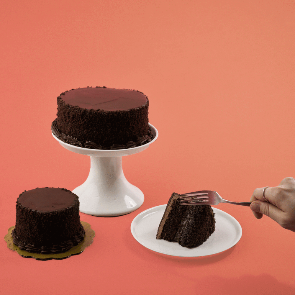 Chocolate cake with chocolate frosting on a white cake stand; a slice of chocolate cake on a white plate with a hand holding a fork; another smaller cake on a gold-colored base, all set against a pink background.