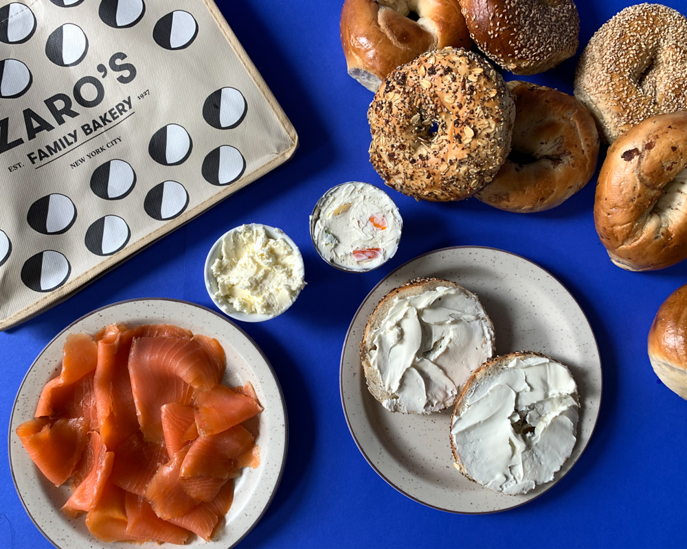 Assorted bagels with cream cheese spreads, smoked salmon, and butter, along with a box from Zarro's bakery on a blue surface.