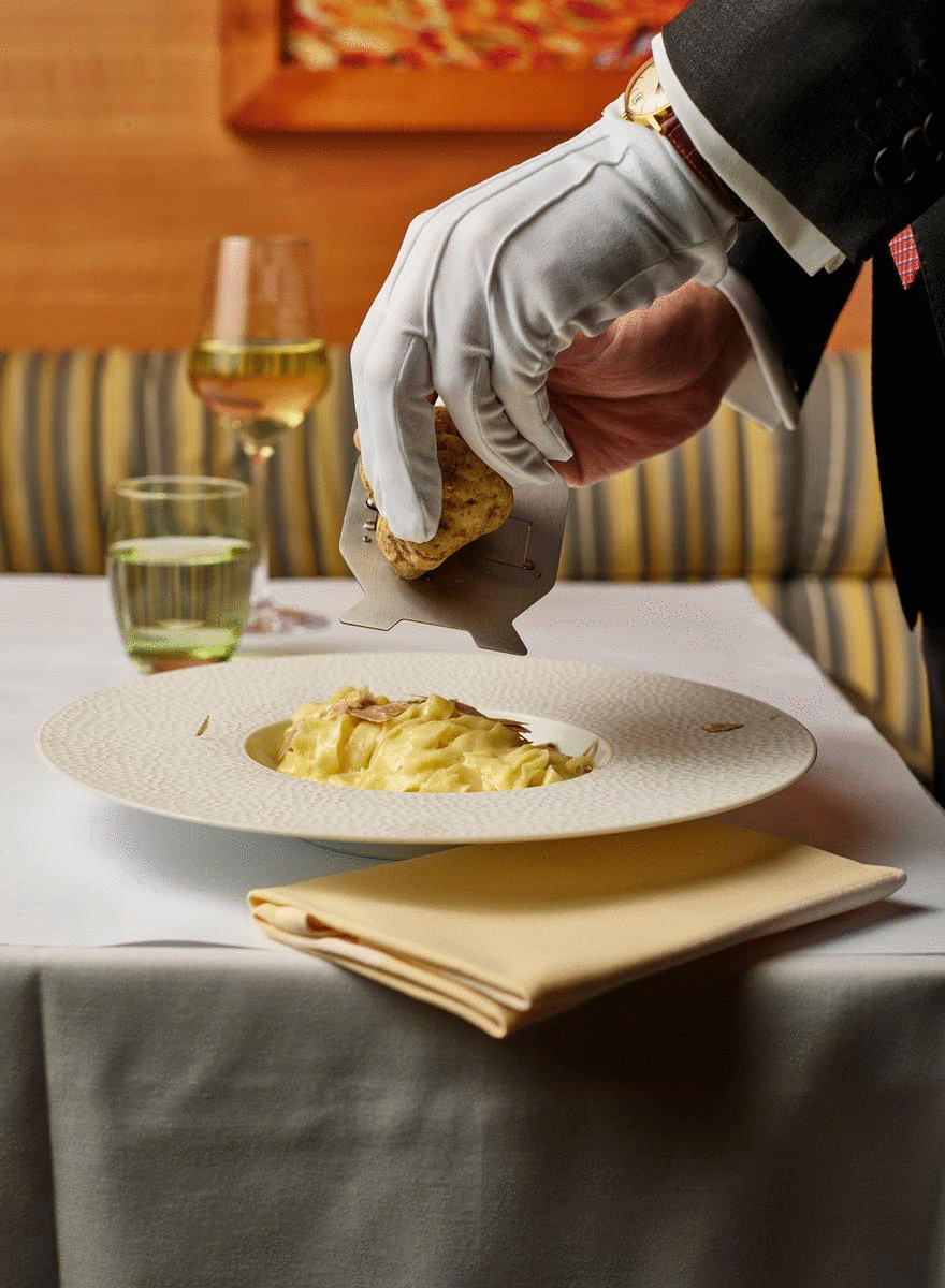 A person in a tuxedo with white gloves seasoning a plate of pasta with grated cheese using a cheese grater, on a table with a glass of white wine, a glass of water, and a napkin.