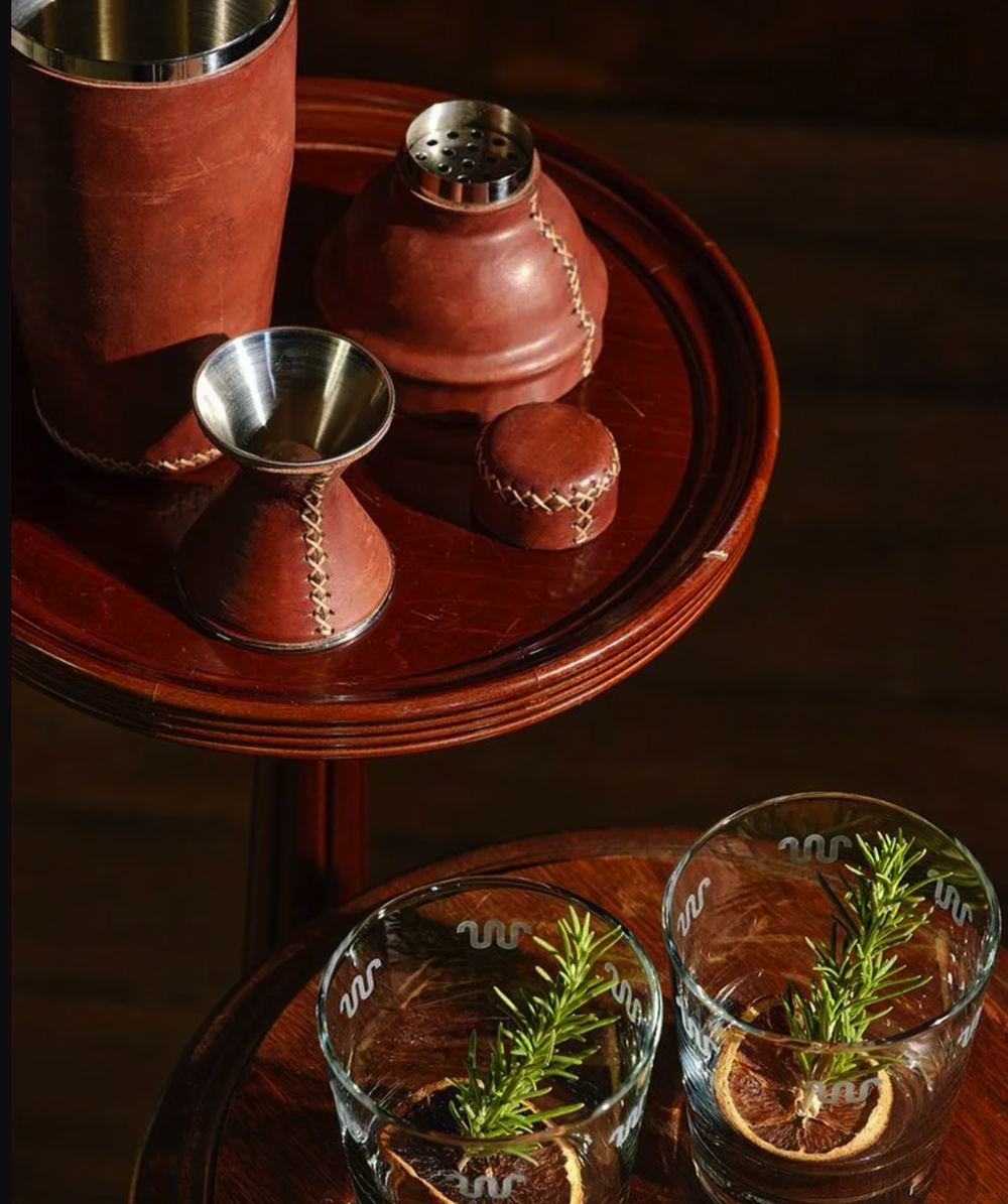 A wooden table holds two glasses with rosemary and lemon slices, and a wooden tray with various leather-covered kitchenware items, including a salt shaker, a small cup, and a larger container.