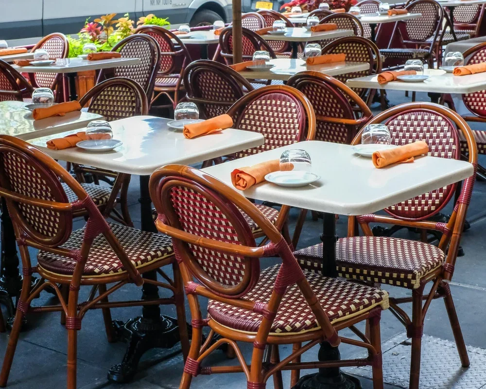 Empty outdoor restaurant with white tables, woven rattan chairs with red and beige checkered upholstery, orange napkins, white plates, and upside-down drinking glasses on each table.