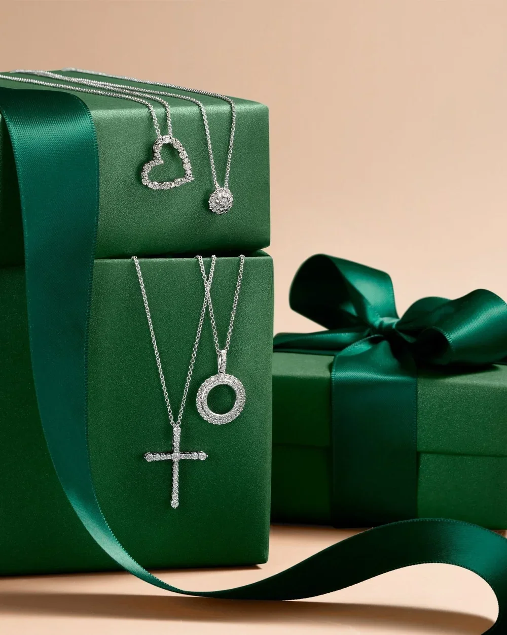 Three silver necklaces with diamond pendants, one in the shape of a heart, one with a circular pendant, and one with a cross, displayed on green gift boxes tied with green satin ribbon.