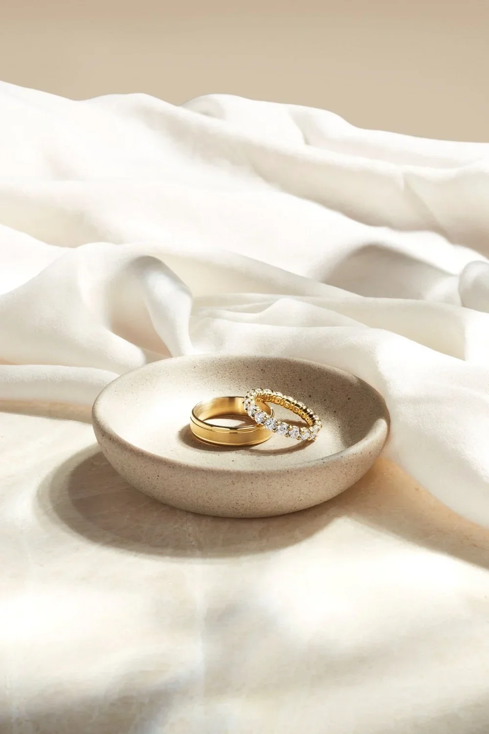 Two gold rings, one plain and one with diamonds, placed in a small beige dish on a white fabric surface.