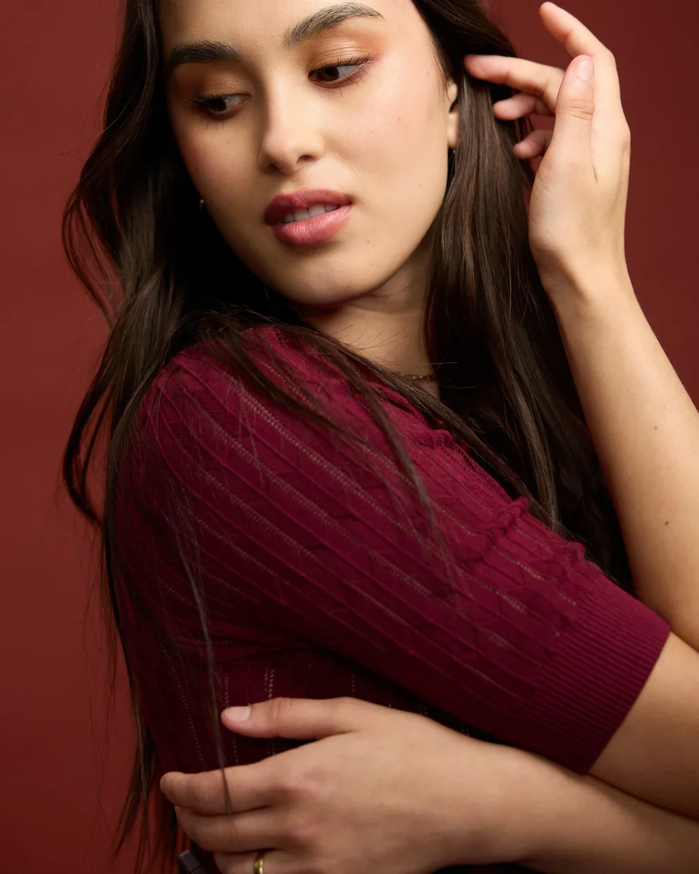 Close-up of a young woman with long dark hair, wearing a burgundy top, looking down with her hand near her ear, against a plain red background.