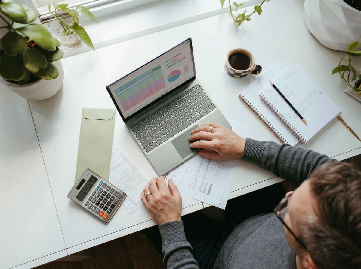 A person on a business valuation at a white desk with a laptop, calculator, cup of coffee, notebooks, and plants.