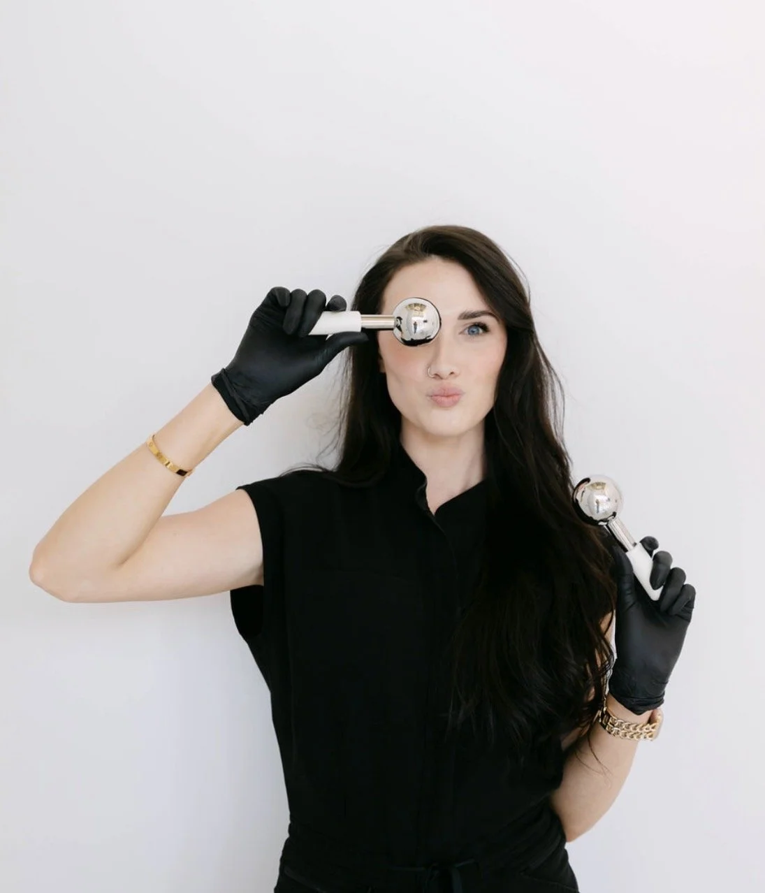 A woman with dark hair holding two silver massage rollers, one on her eye and the other near her shoulder, wearing black gloves, a black shirt, gold watch, and a bracelet, standing against a white wall, making a playful facial expression.