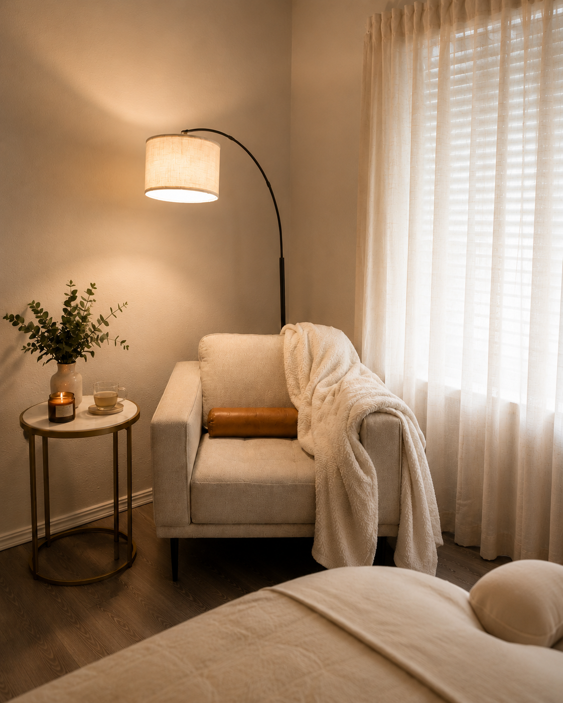 Living room corner with a beige armchair, a blanket, a pillow, a side table with a plant, candle, mug, and a small decorative object, a standing lamp, and sheer curtains.