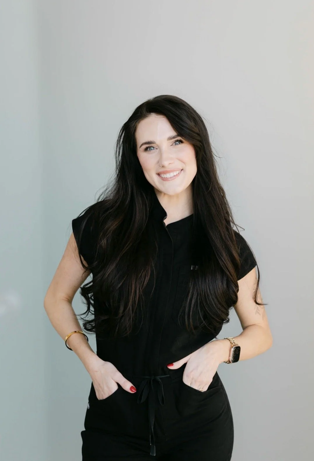 A woman with long, wavy black hair and light skin smiling, wearing a black sleeveless top, black pants with a drawstring, a gold bracelet, and a smartwatch, standing against a neutral background.
