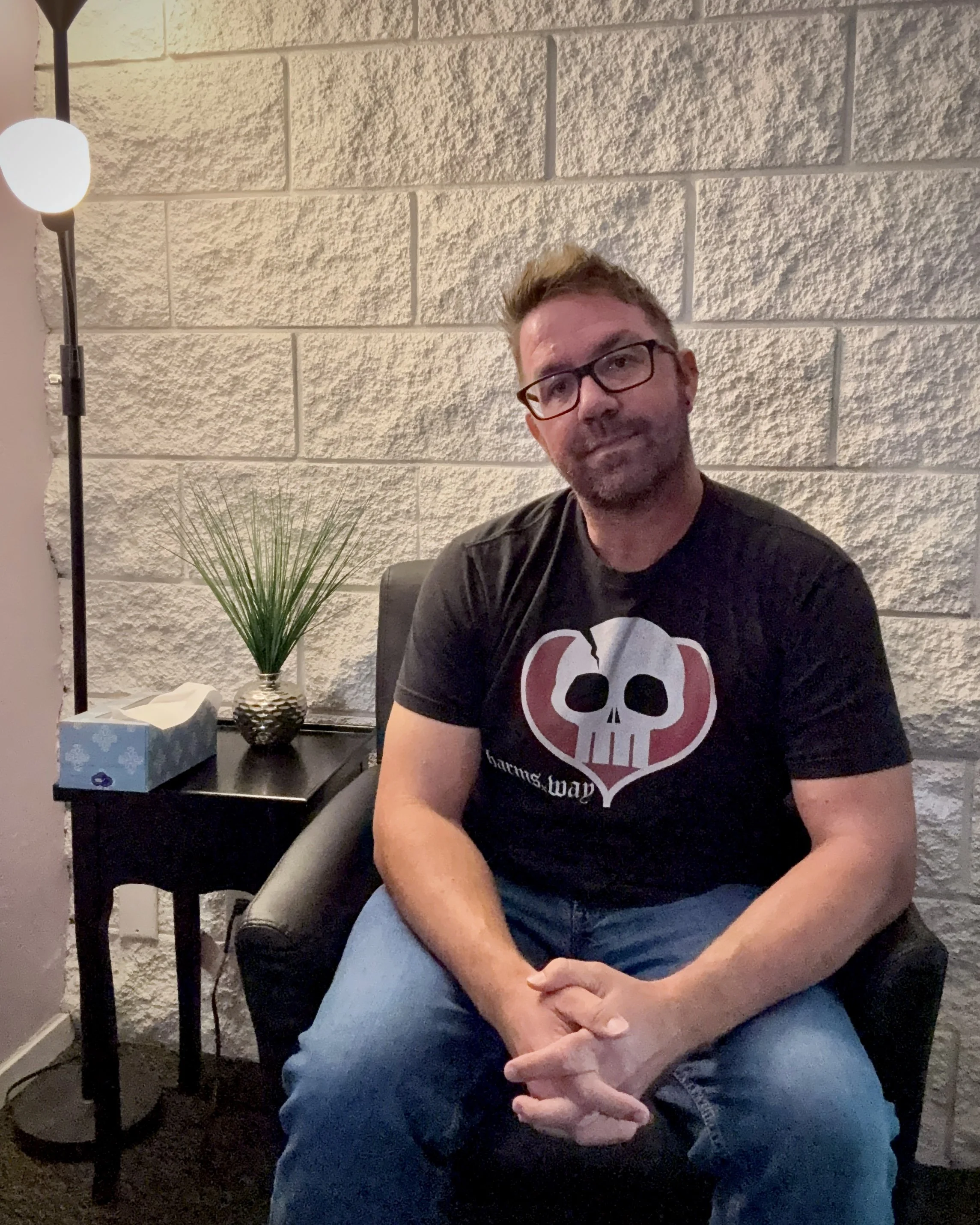A man wearing glasses and a black t-shirt with a white skull graphic is sitting in a chair with his arms crossed, in front of a textured beige brick wall. A table to his right holds a tissue box and a decorative vase with green grass. A tall lamp with a round white shade and black stand is on his left.