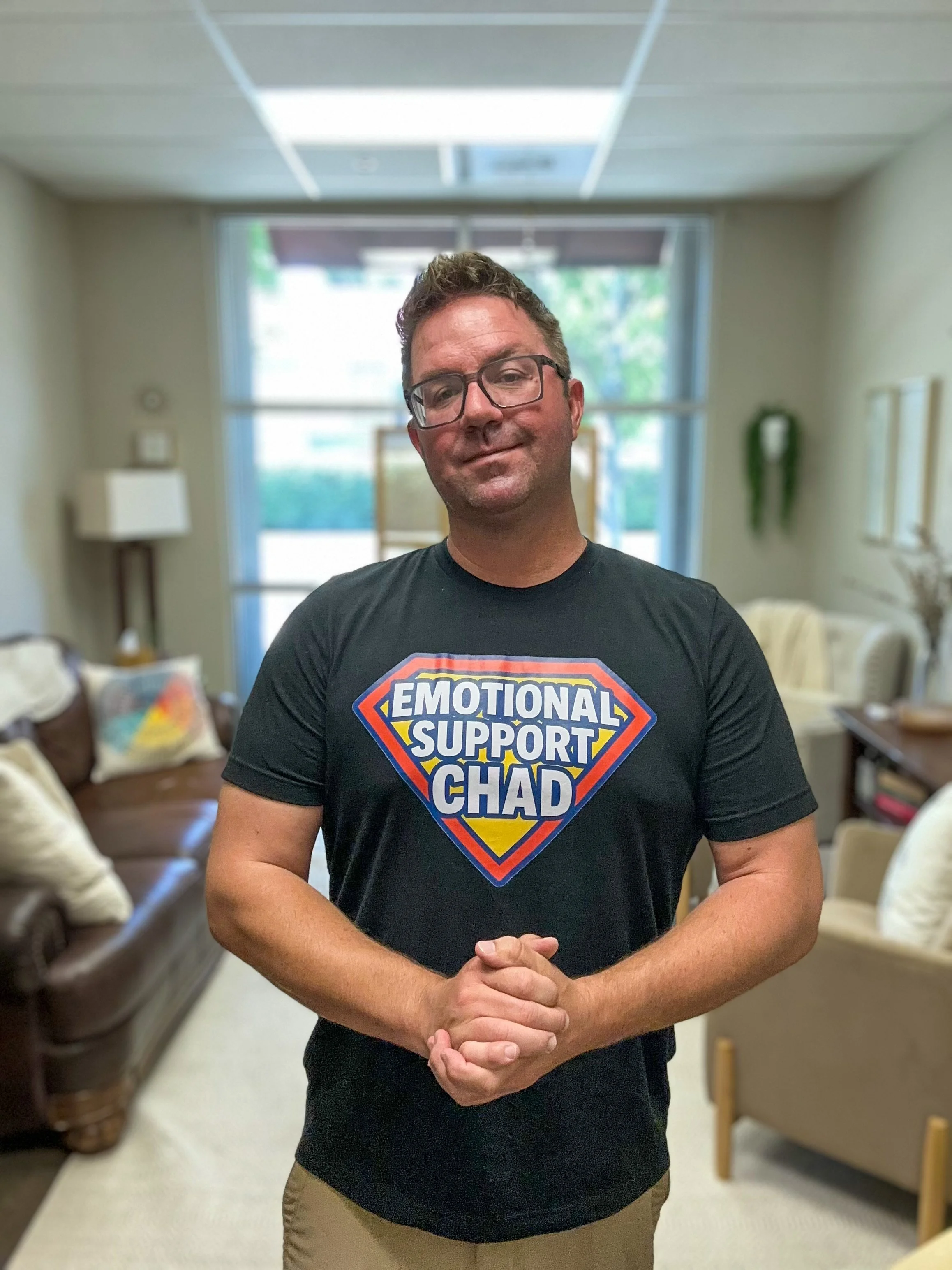 A man wearing glasses and a black T-shirt with a logo that reads 'Emotional Support Chad' in a room with beige walls, chairs, and a window in the background.