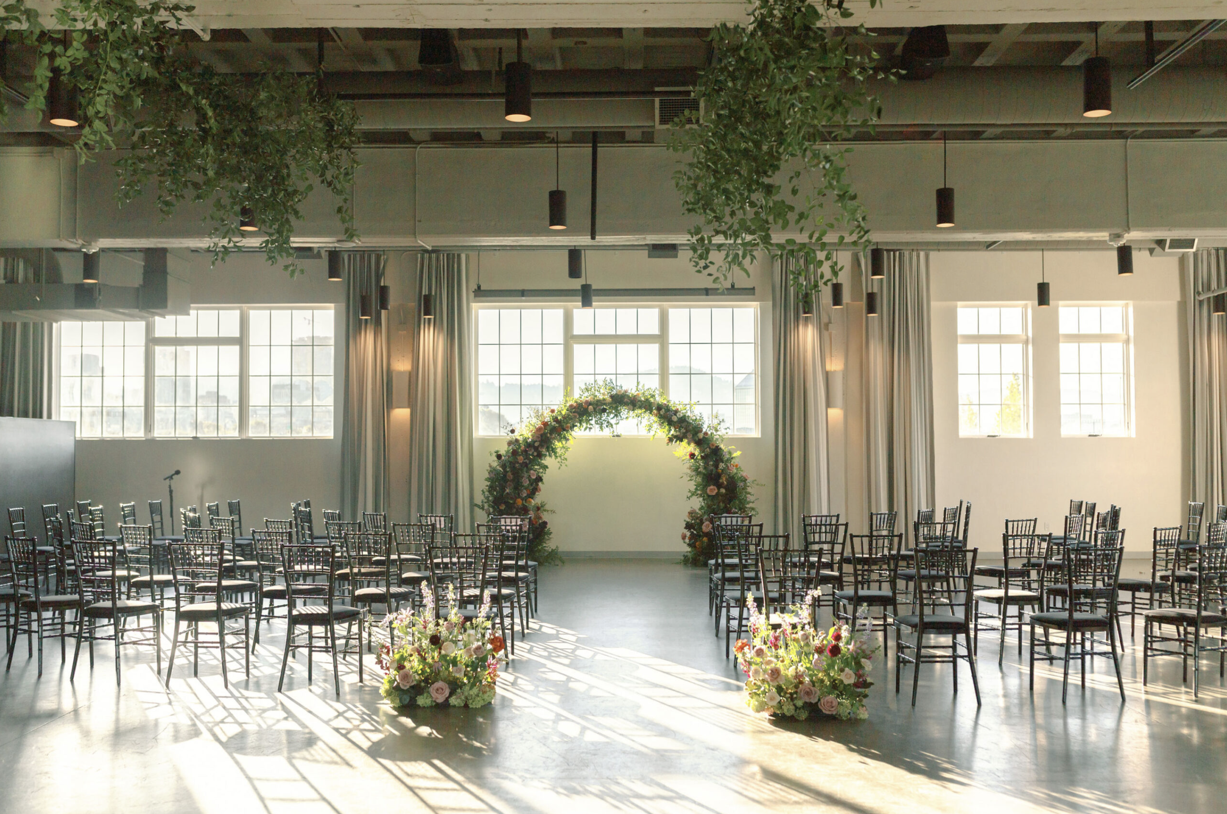 Wedding ceremony setup inside The Exchange with rows of black chairs, floral arrangements, and large windows.