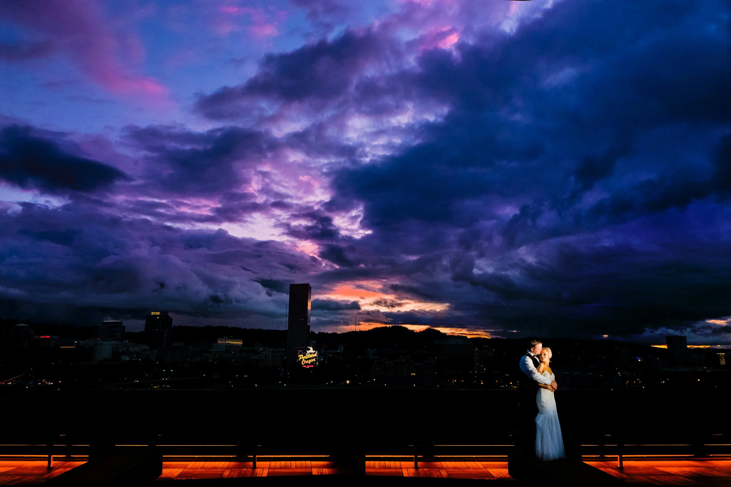 A couple dressed in wedding attire embracing each other on a rooftop at sunset, with a city skyline and dramatic dark purple and pink cloud-filled sky in the background.