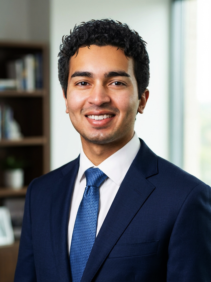 A smiling man in a navy blue suit, white shirt, and blue tie, standing in an office with bookshelves and a plant in the background.