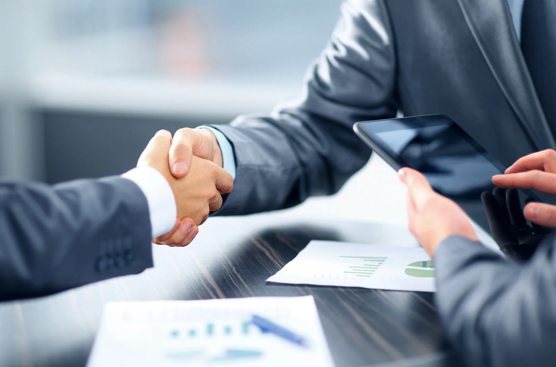 Two people in suits shaking hands across a table, with one person holding a tablet, in a business meeting setting.
