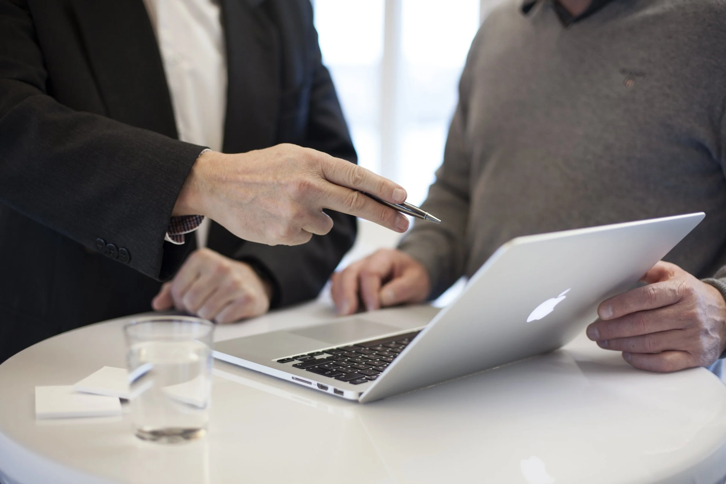 Two men in business attire discussing while looking at a laptop and holding a pen, with coffee on a white table.