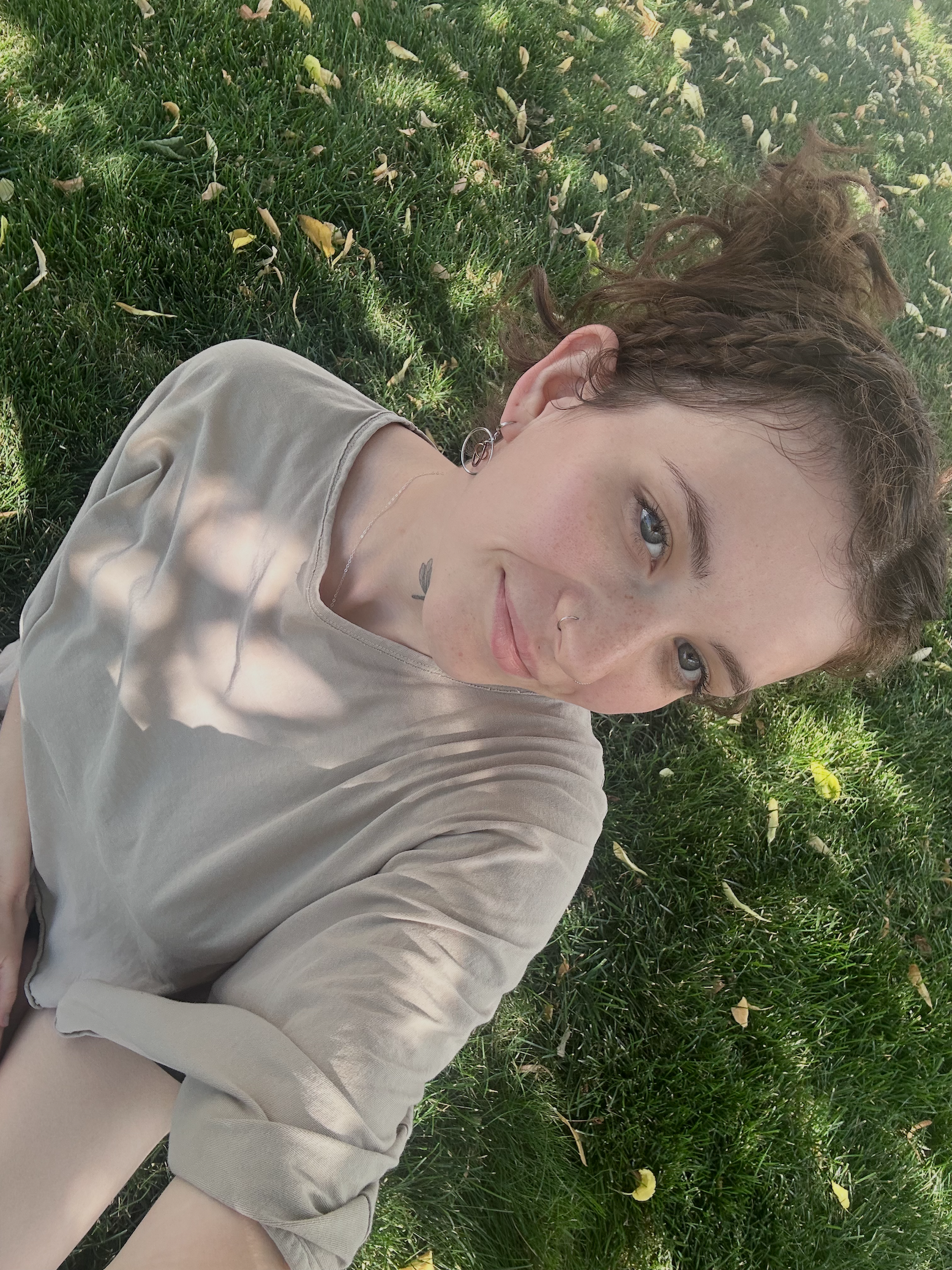 A woman with curly hair sitting on grass outside, wearing a beige shirt, with a small tattoo on her neck and earrings, smiling and looking at the camera.