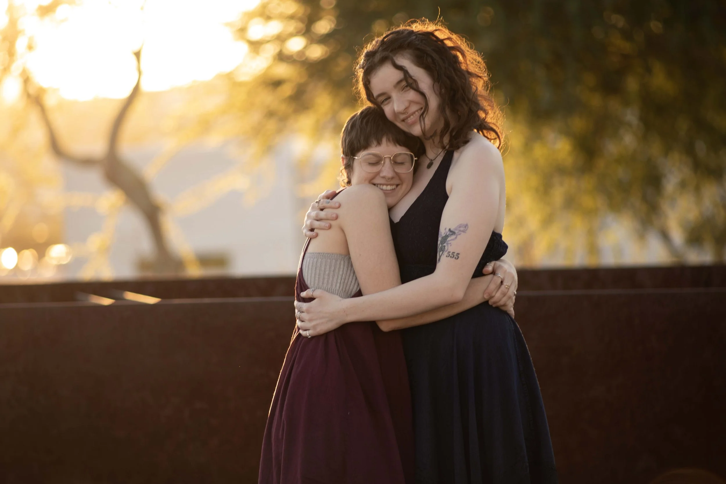 Two people embracing outdoors during sunset, smiling with eyes closed, warm light illuminating their faces, trees in background.