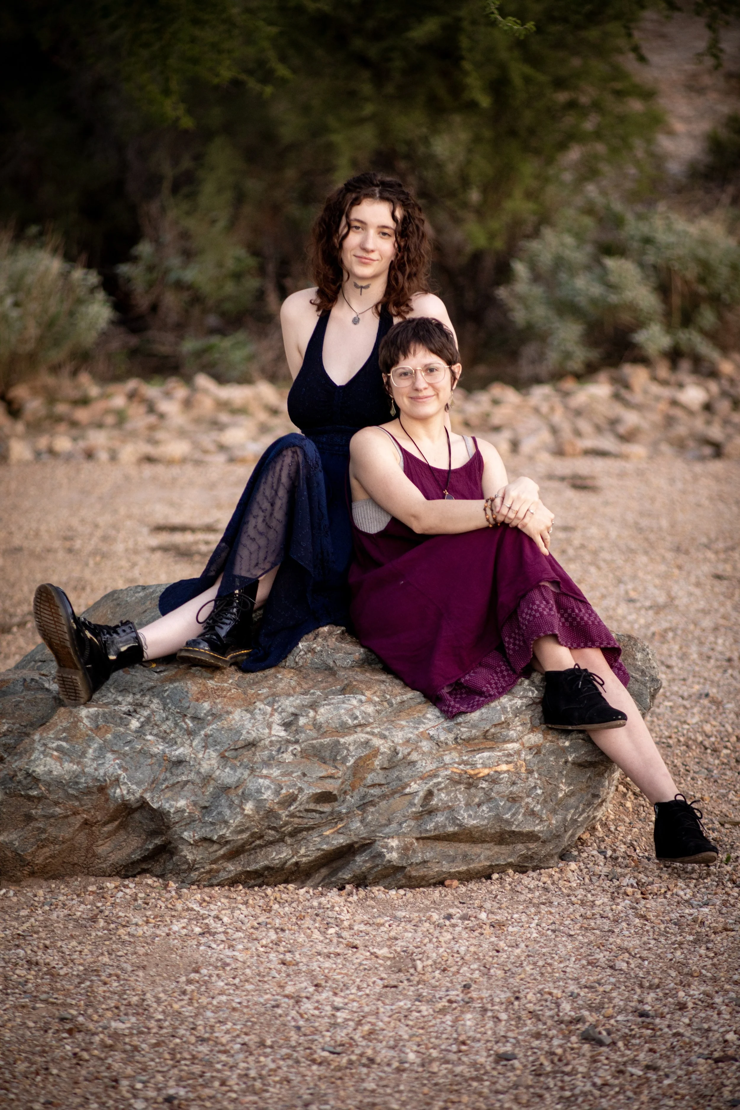 Two young people. One with short hair, one with curly hair. Sitting on a large rock outdoors, with trees and gravel in the background.