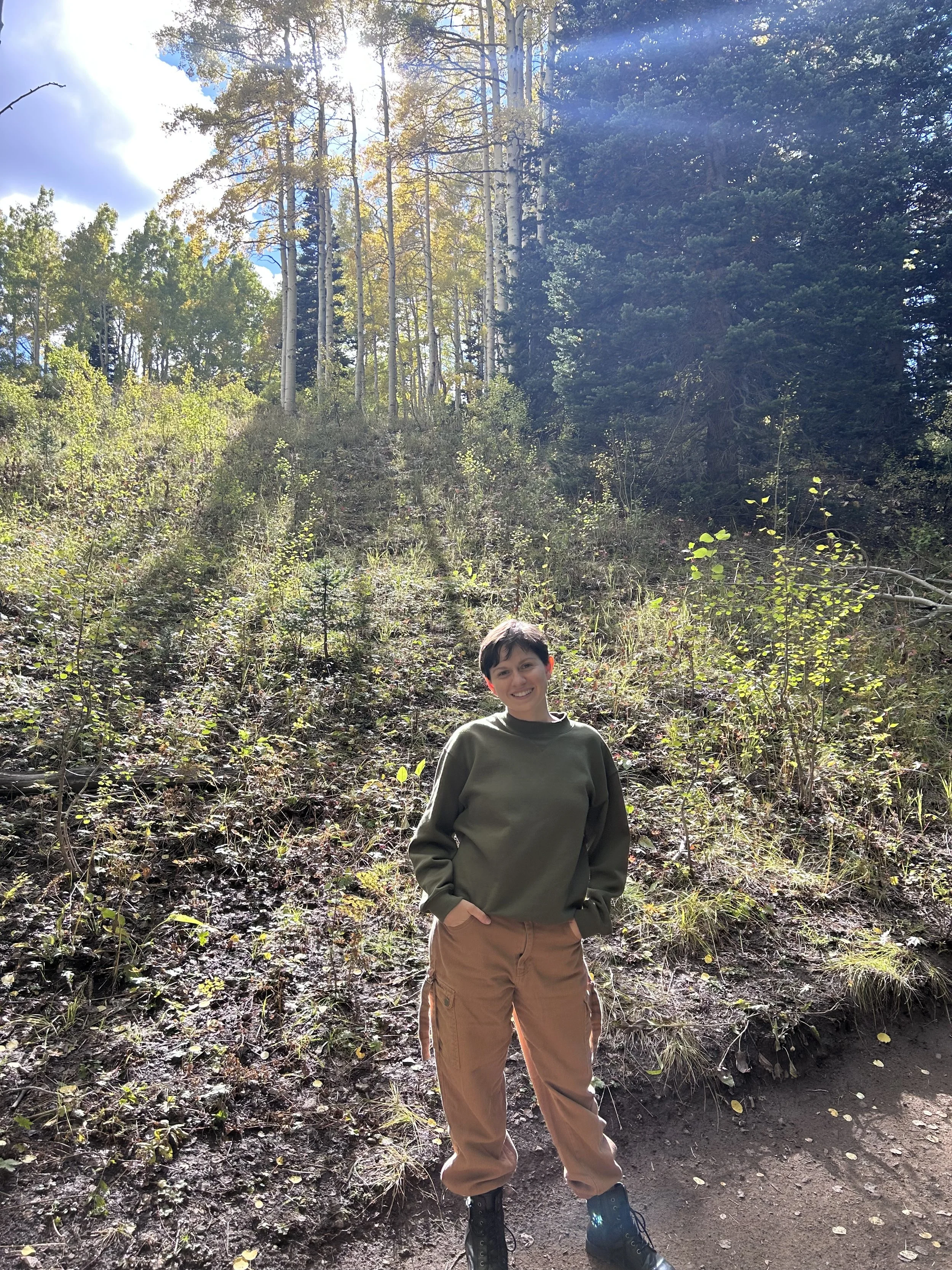 A person standing outdoors in a forested area with sunlight streaming through the trees, smiling and looking at the camera.