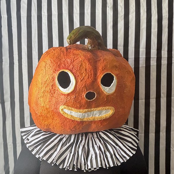 A pumpkin painted with a face, featuring large eyes, a small nose, and a smiling mouth, placed on a striped cloth with a striped background.