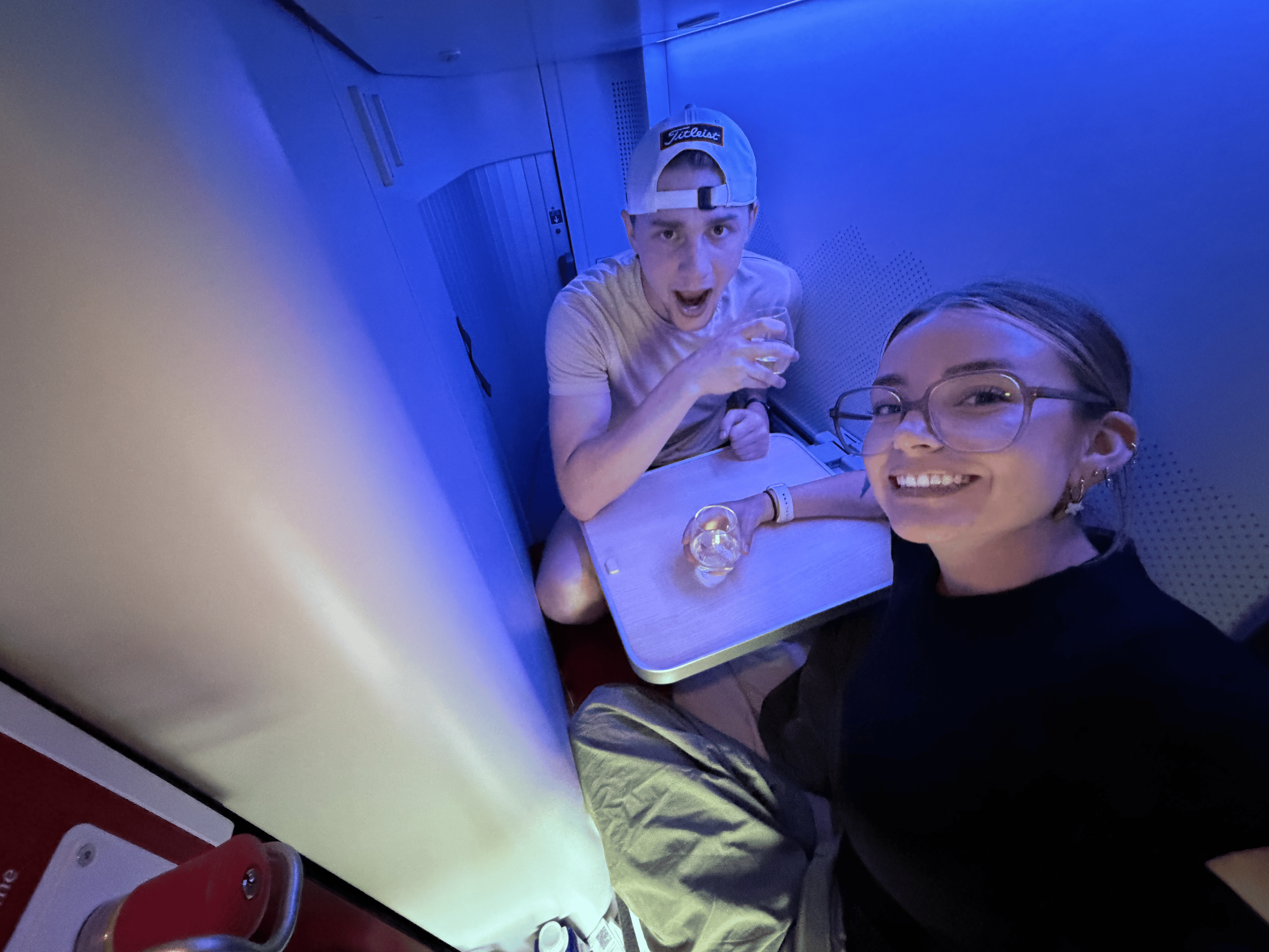 Two people are taking a selfie on a train or bus. The young man is wearing a baseball cap backwards and has a surprised expression while holding a drink. The young woman, with glasses, is smiling. There is a small table with a glass on it, and the background shows metallic panels and seating.