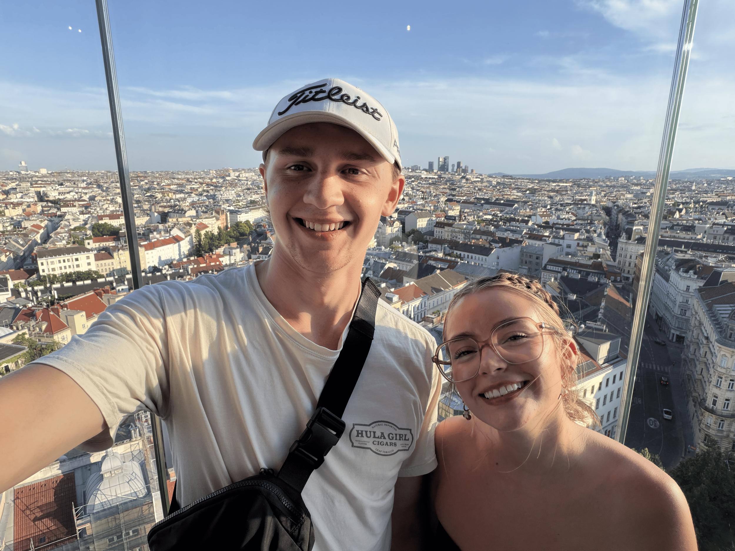 Two smiling young adults, a man and a woman, taking a selfie atop a tall structure with a cityscape in the background. The man wears a white cap with 'Titleist' written on it, and the woman wears glasses. The city below features numerous buildings and a clear blue sky.