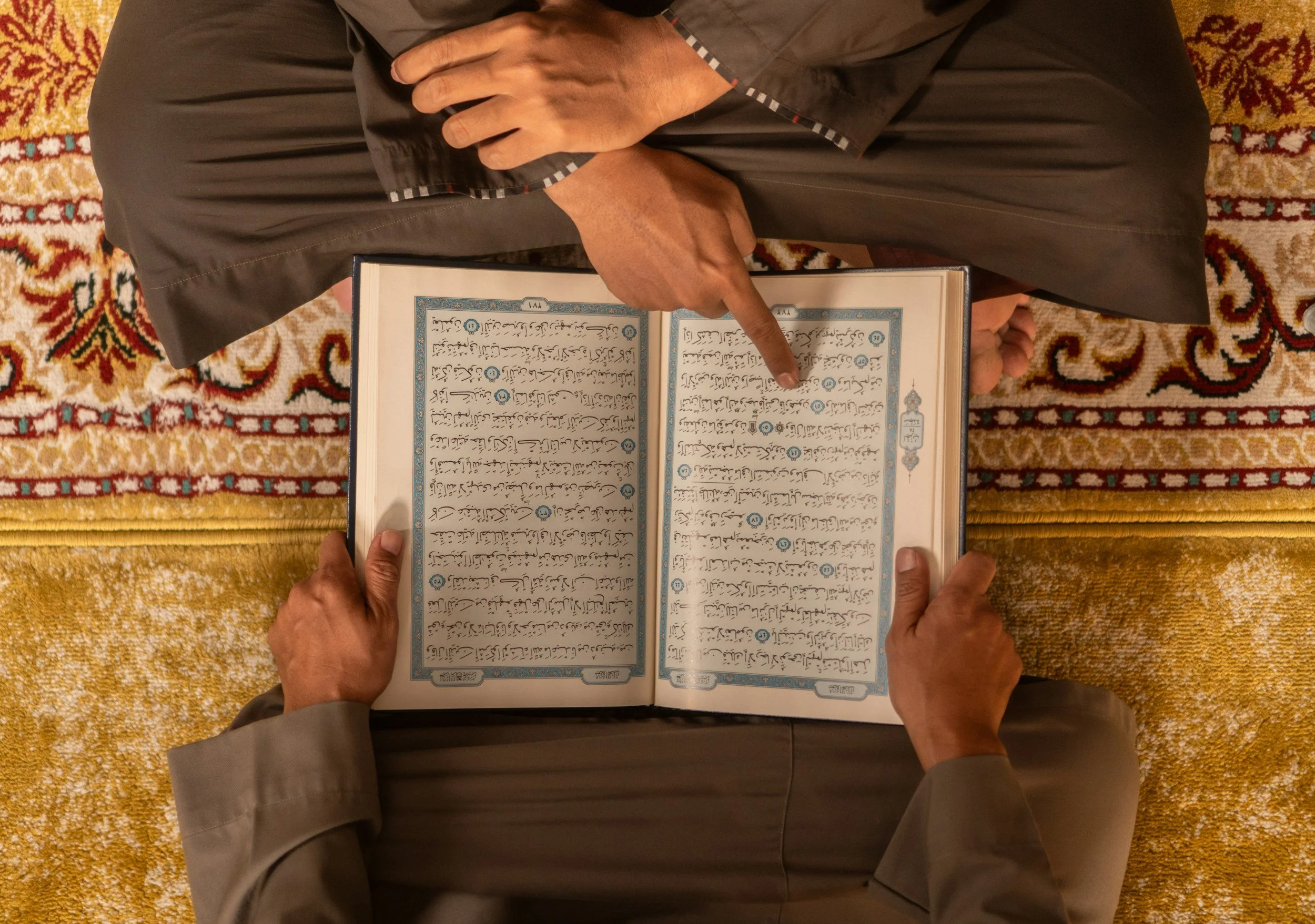 A person dressed in a dark gray robe or suit is sitting on a patterned carpet, holding an open book with handwritten script. The person is using one hand to hold the book and is pointing at the text with the index finger of their other hand.