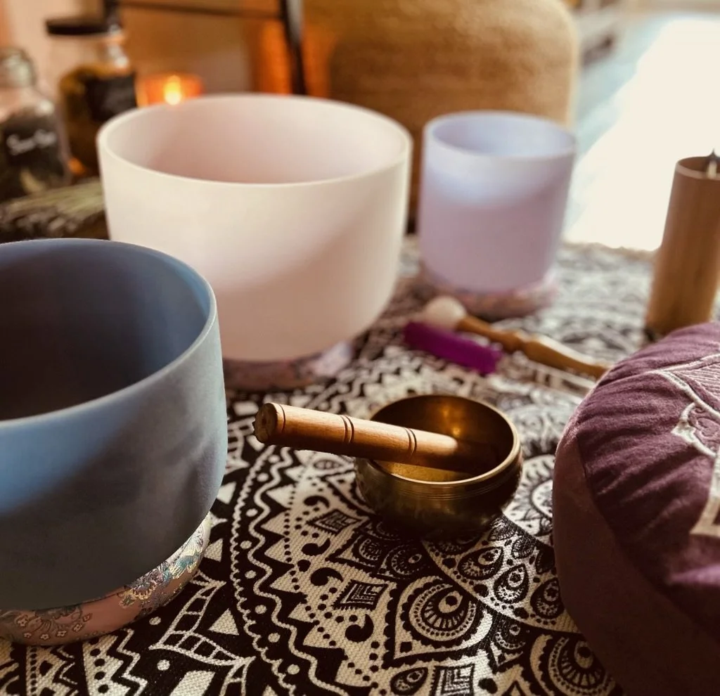 Several singing bowls of different sizes and colors on a patterned cloth, with a wooden mallet resting inside a metallic bowl, on a table with dim lighting and candles in the background.