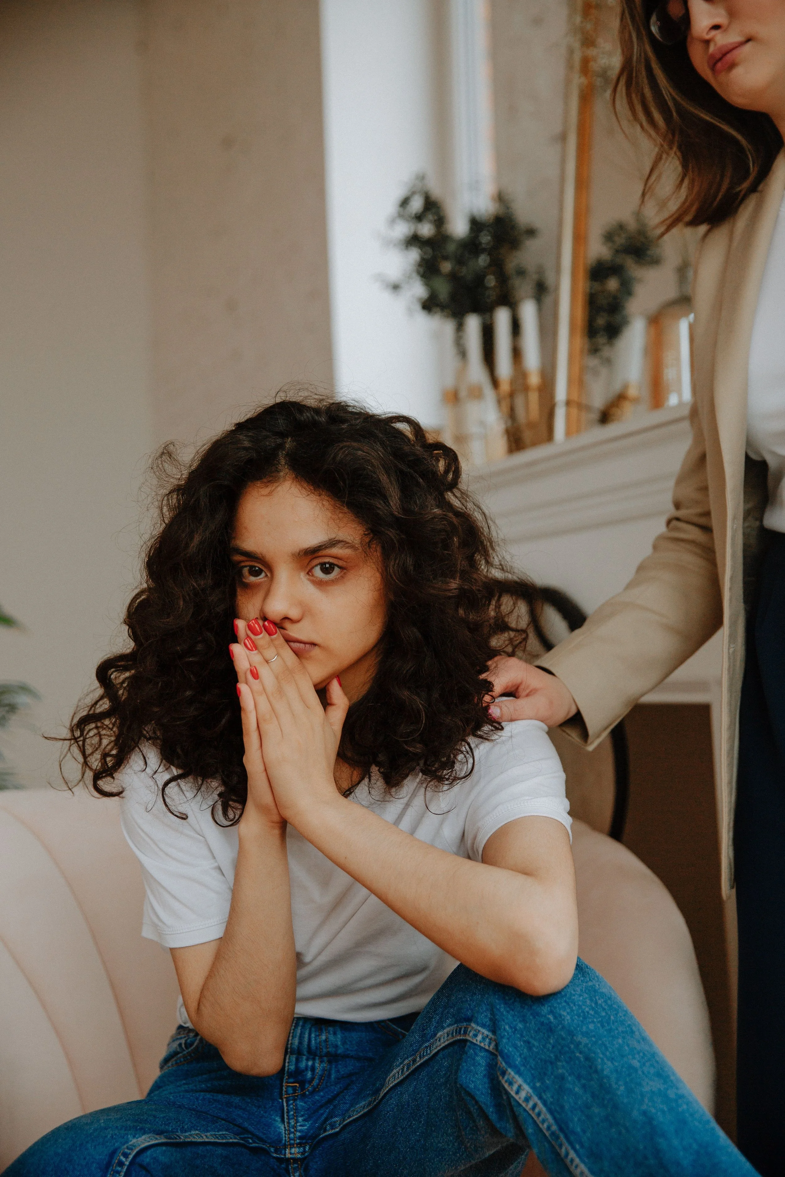 A young woman with dark, curly hair and light skin sitting on a beige chair, looking directly at the camera, with her hands pressed together near her face, wearing a white t-shirt and blue jeans. Another woman with light skin, brown hair, glasses, and a beige blazer is standing beside her, touching her shoulder. The background features a white wall, a window, a mirror, and some potted plants.