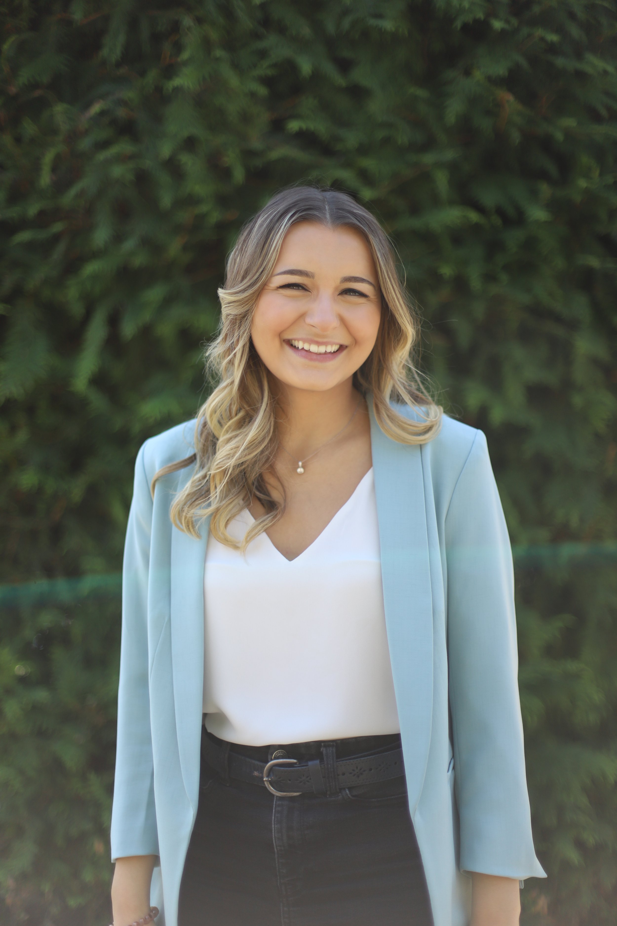 A woman with blonde hair styled in loose waves, smiling outdoors in front of green foliage, wearing a light blue blazer over a white top and black pants.