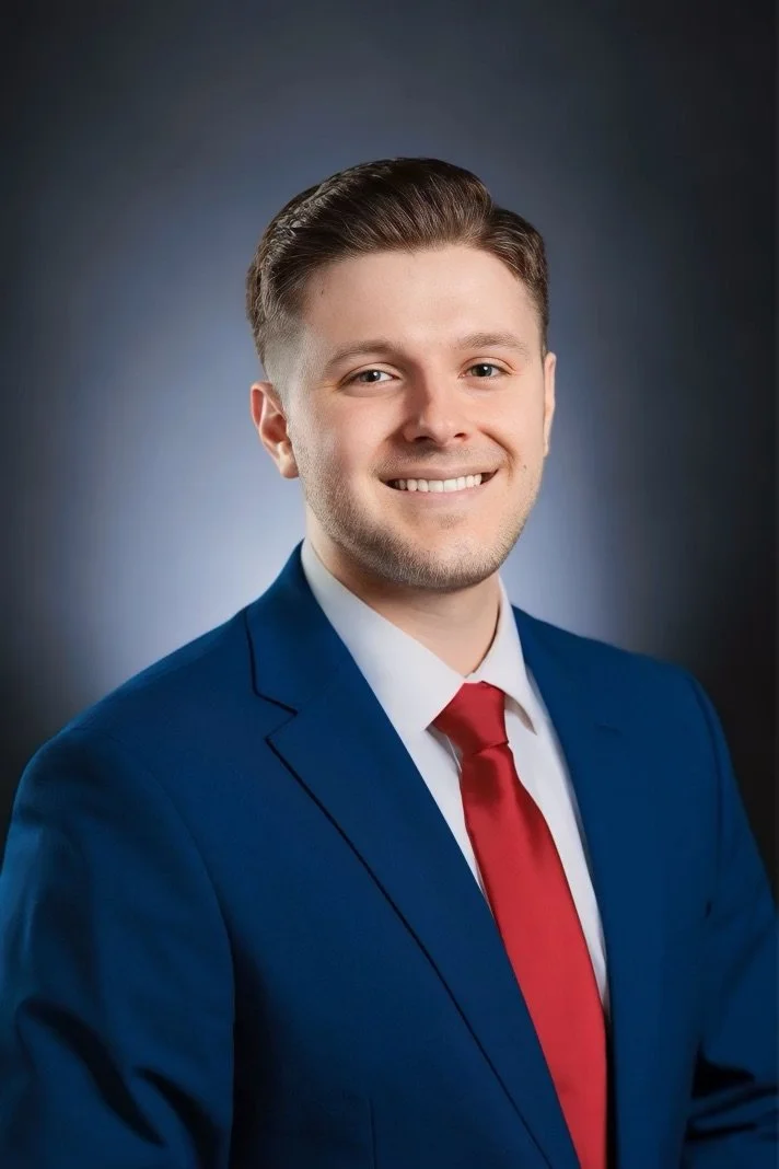 Young man with short brown hair, wearing a blue suit, white shirt, and red tie, smiling in a professional portrait.