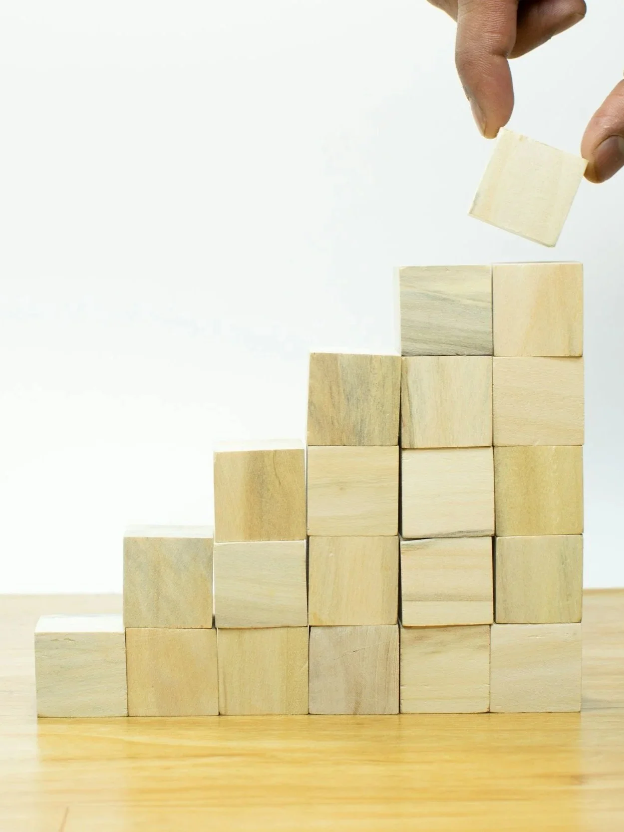 A hand placing a wooden block on top of a stack of wooden blocks arranged in a staircase pattern.